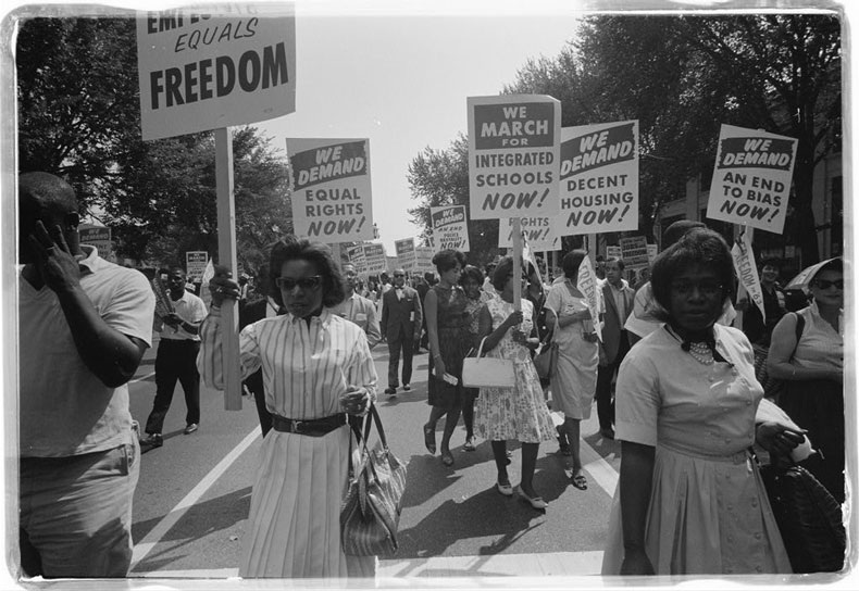 Photograph shows a procession of African Americans carrying signs for equal rights, integrated schools, decent housing, and an end to bias.